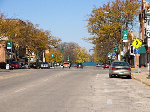 A quiet street lined with trees and shops, featuring cars parked along the side and a view of a lake in the distance.