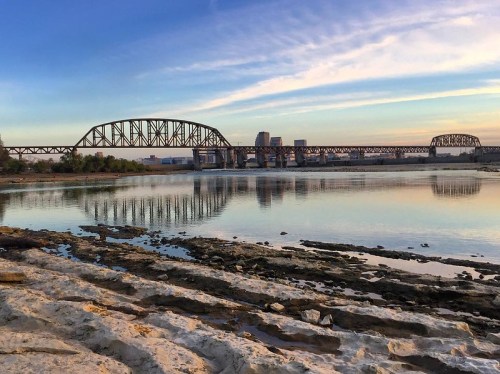 A scenic view of a river with a bridge in the background, reflecting the sky and surrounded by rocky shorelines.