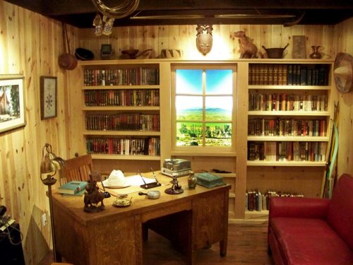 Cozy study with wooden walls, bookshelves filled with books, a desk, and a window showing a scenic view.