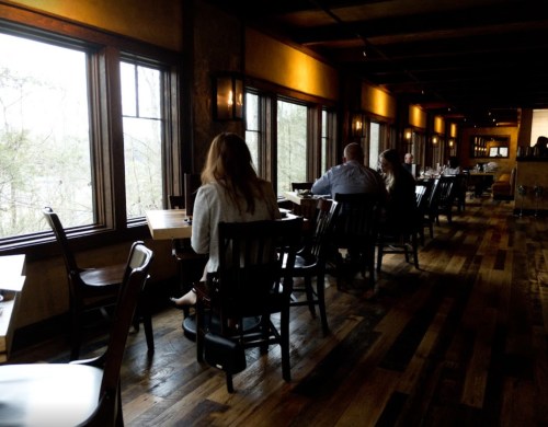 A cozy restaurant interior with wooden floors, tables, and patrons seated by large windows overlooking nature.