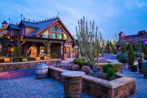A charming rustic building with a landscaped courtyard, surrounded by trees and barrels, under a twilight sky.