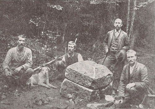 Five men and a dog pose around a stone well in a wooded area, dressed in late 19th-century attire.