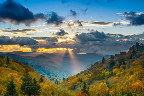 A scenic view of mountains at sunset, with rays of light breaking through clouds over colorful autumn foliage.