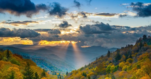 A scenic view of mountains at sunset, with rays of light breaking through clouds and colorful autumn foliage.
