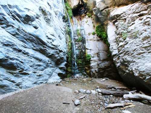 A serene waterfall cascading down rocky cliffs, surrounded by lush greenery and a pebbled ground.