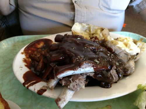 A plate of barbecued ribs covered in sauce, served with a baked potato and a side of coleslaw.