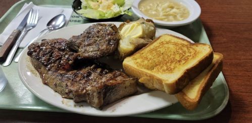 A plate with grilled steak, baked potato, salad, soup, and two slices of toasted bread on a tray.