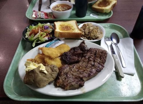 A tray with a steak, baked potato, salad, fried fish, toast, and a side of mushrooms, with utensils and condiments.