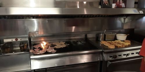 A grill with sizzling meat and toast, with a small flame on the grill in a stainless steel kitchen.