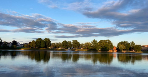 A serene lake reflecting trees and clouds under a blue sky, with a small building visible on the shore.
