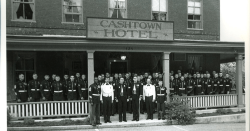 A large group of military personnel poses in front of the Cashtown Hotel, with a historic building in the background.