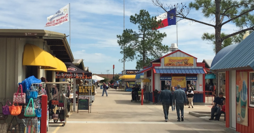 A vibrant outdoor market with colorful shops, flags, and visitors walking along a paved path under a blue sky.