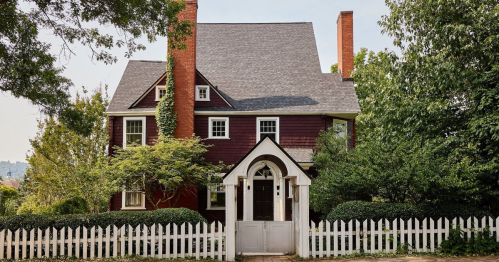 A charming red house with a steep roof, white trim, and a picket fence, surrounded by lush greenery.