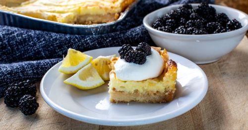 A slice of cheesecake topped with blackberries and whipped cream, served with lemon wedges, alongside a bowl of blackberries.
