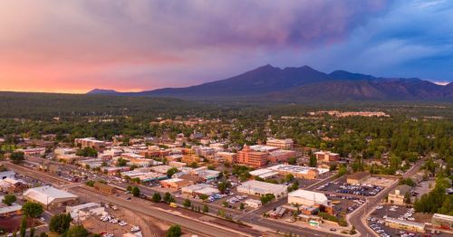 Aerial view of a town surrounded by trees and mountains at sunset, with colorful skies and buildings visible below.