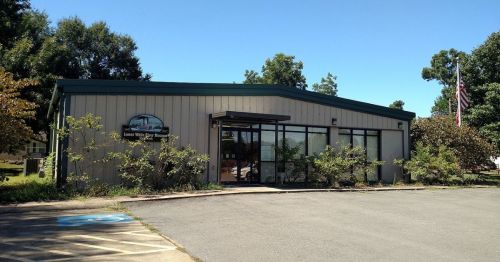 A single-story building with a green roof, large windows, and surrounding trees, featuring a sign and American flag.