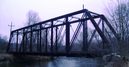 A dark, metal truss bridge spans a river, surrounded by bare trees and a cloudy sky.