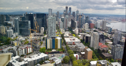 Aerial view of a city skyline featuring tall buildings, greenery, and a waterfront under a cloudy sky.