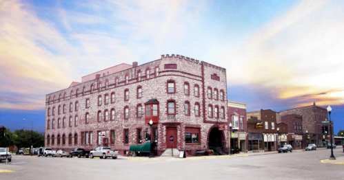 Historic brick building at a street corner, with shops and vehicles, under a colorful sunset sky.