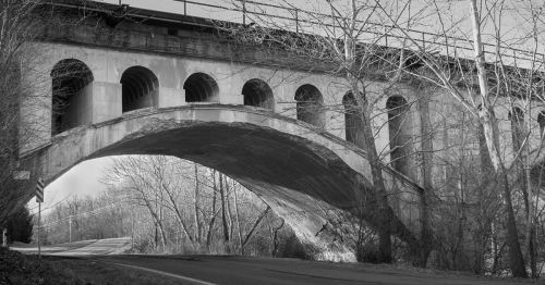 Black and white image of a large arch bridge with multiple arches, surrounded by bare trees and a winding road below.