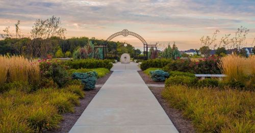 A serene garden path leads to an archway, surrounded by lush greenery and colorful plants under a cloudy sky.