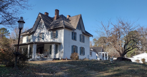 A charming two-story white house with a steep roof, surrounded by trees and a grassy yard on a clear day.