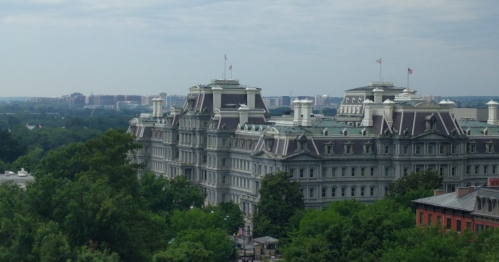 A historic building with a gray facade and multiple flags, surrounded by green trees and a distant city skyline.