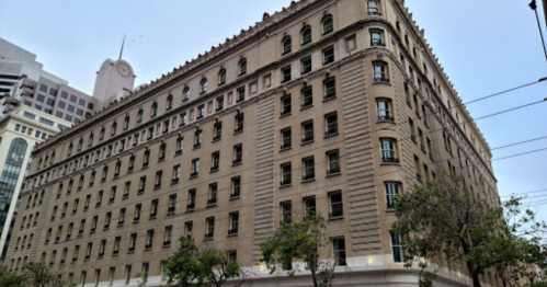 Historic building with a beige facade, multiple windows, and a decorative roof, surrounded by trees and cityscape.