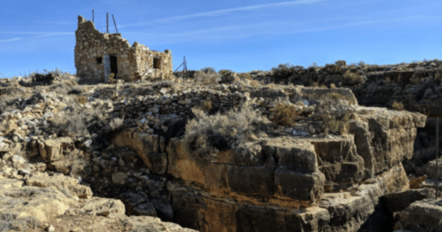 Ruins of a stone building on rocky terrain, surrounded by sparse vegetation under a clear blue sky.