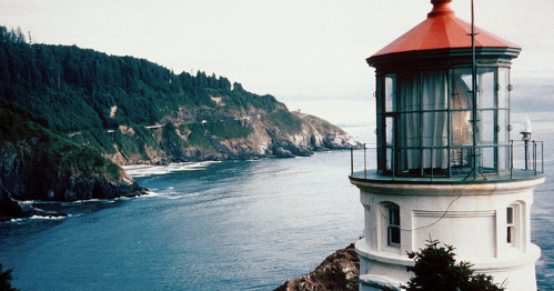 A coastal lighthouse overlooking a rocky shoreline and calm ocean, surrounded by lush green hills.