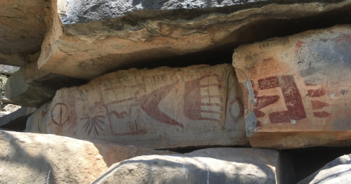 Ancient rock art featuring red and ochre symbols on a stone surface, partially shaded by surrounding rocks.