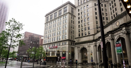 A rainy city street featuring tall buildings, traffic lights, and a bus stop, with greenery in the foreground.