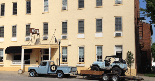 A light blue truck with a trailer carrying a vintage jeep parked in front of a yellow hotel building.