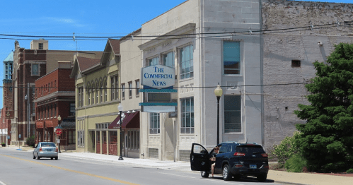 A street view featuring a building with "The Commercial News" sign, parked cars, and clear blue skies.