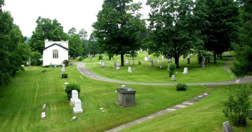 A serene cemetery with green grass, trees, and a small chapel surrounded by gravestones along winding paths.