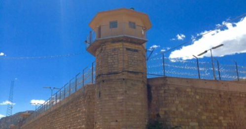 A tall watchtower atop a stone prison wall, surrounded by barbed wire under a bright blue sky.