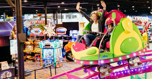 A woman joyfully rides a colorful amusement park ride with her hands raised, surrounded by arcade games.