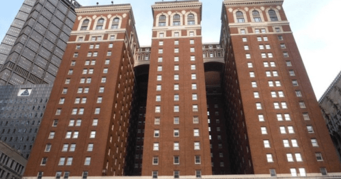 Three tall, red-brick buildings with arched openings, set against a blue sky and surrounded by modern architecture.