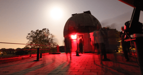 A dimly lit observatory at night, with people moving around under a bright moon and red lights illuminating the scene.