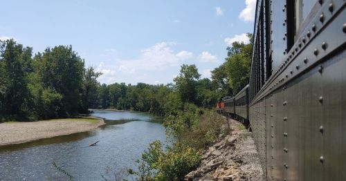 A train travels alongside a river, surrounded by lush greenery and a clear blue sky.