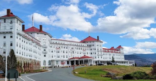 A large white hotel with red roofs, set against a blue sky and mountains, surrounded by green grass and trees.