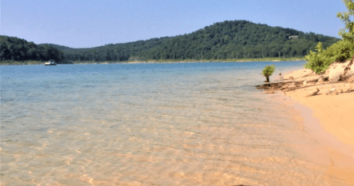 A serene lake scene with clear water, sandy shore, and green hills in the background under a blue sky.