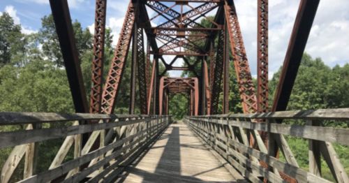 A weathered metal bridge with wooden planks, surrounded by lush greenery and a blue sky.