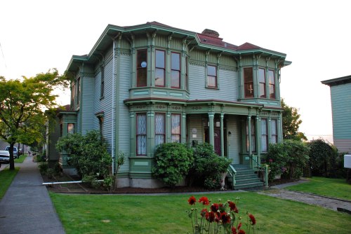A large, green Victorian-style house with ornate details, surrounded by lush greenery and flowers in the foreground.