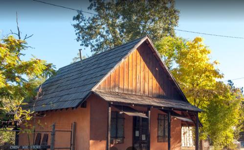 A small, rustic building with a wooden roof and earthy walls, surrounded by trees in a sunny setting.