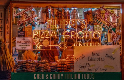 A vibrant storefront of Flippi's Pizza Grotto, featuring neon signs and a menu of Italian foods, including cannoli shells.