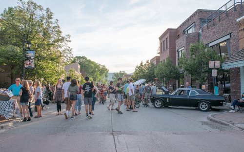 A lively street scene with a crowd of people, bicycles, and a classic car, surrounded by shops and trees.
