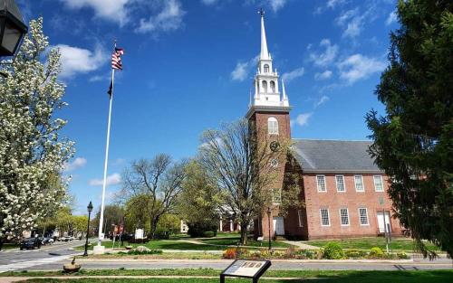 A brick church with a tall steeple, surrounded by trees and a flagpole, under a blue sky with fluffy clouds.