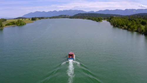 A boat glides through a calm river, surrounded by lush greenery and distant mountains under a clear blue sky.