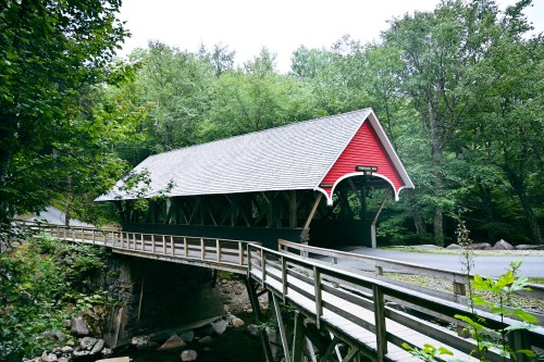 A red covered bridge spans a small stream, surrounded by lush green trees and a gravel road.